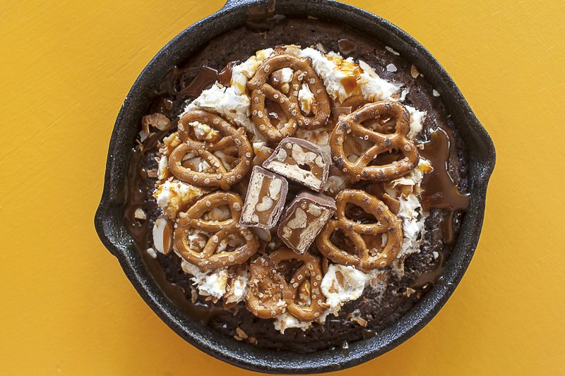 Brownie Dessert Pan Pizzas in a cast iron skillet with pretzels, mini candy bars and chocolate drizzle
