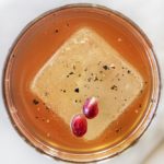 One large ice cube floating in a whisky cocktail with pomegranate seeds and black pepper