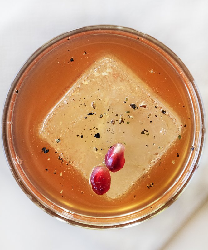 One large ice cube floating in a whisky cocktail with pomegranate seeds and black pepper