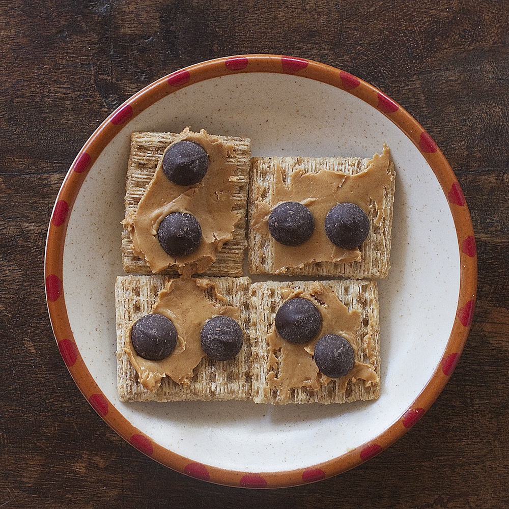 Four Triscuits arranged on a small plate. Each is spread with creamy peanut butter and has two chocolate chips pressed into