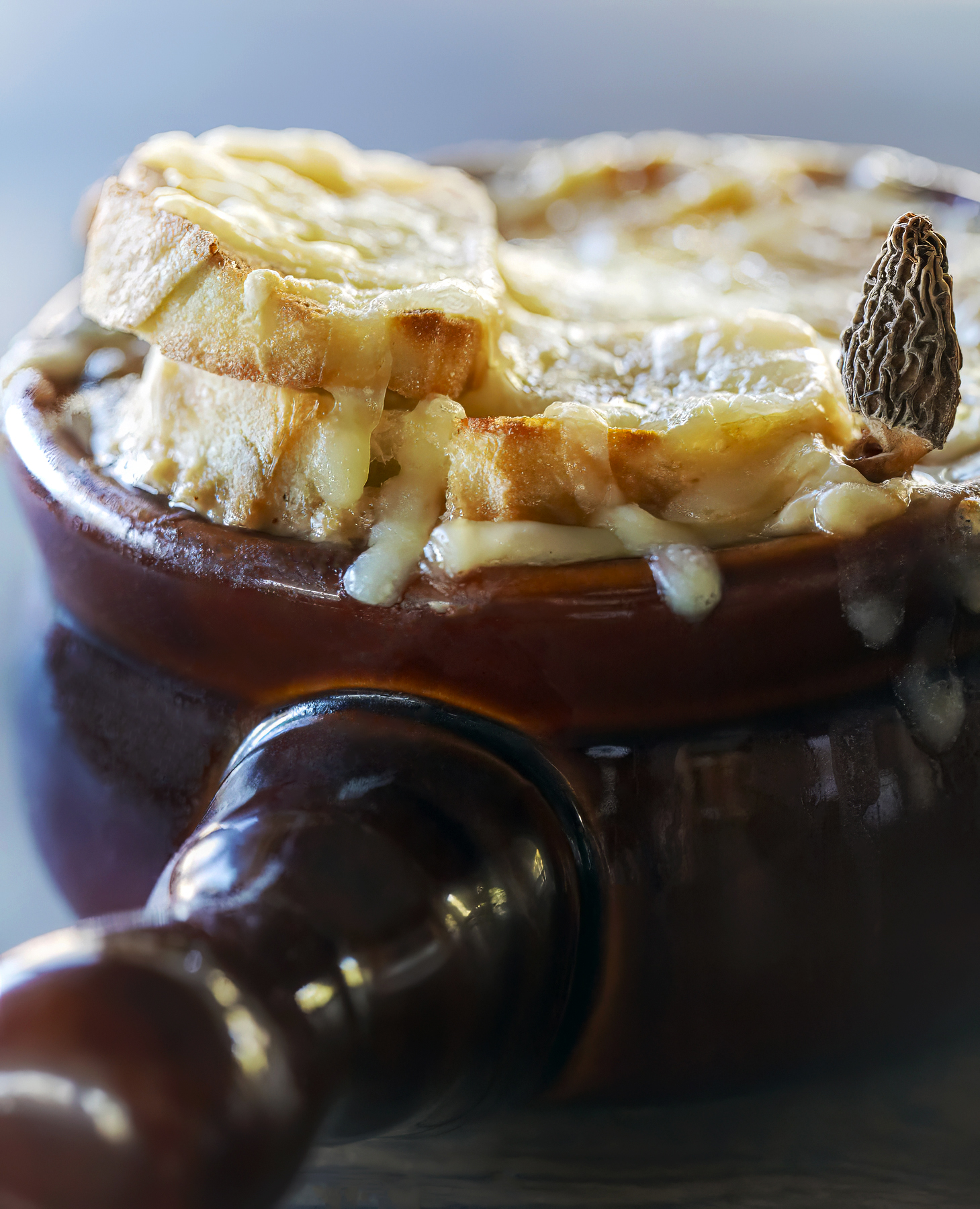 Soup tureen filled with wild morel mushroom and onion, acorn soup and topped with cheese, a dried morel mushroom and toasted baguette slices.